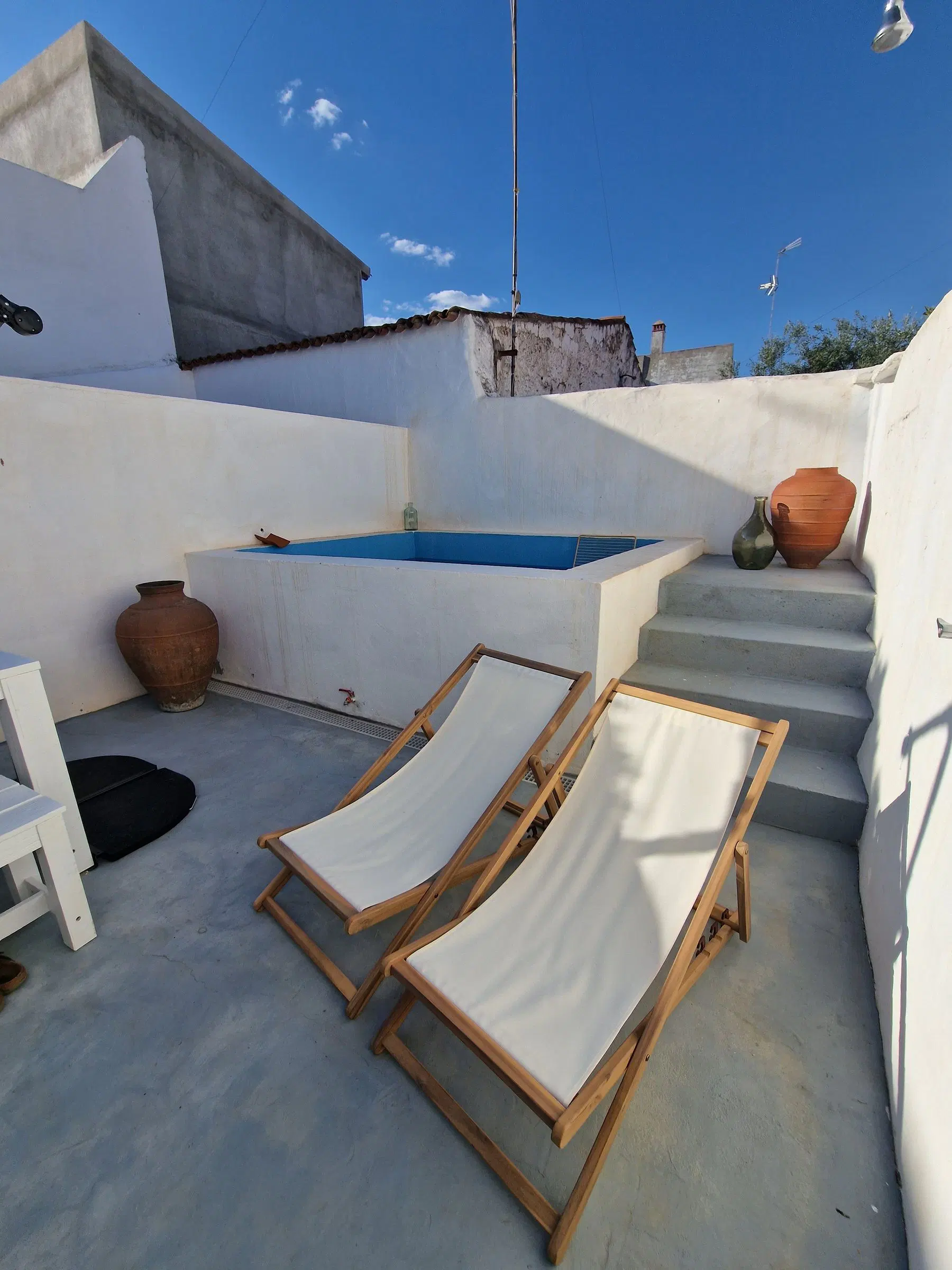 Piscina retangular encastrada no pátio caiado da casa, com duas espreguiçadeiras de lona branca e madeira, uma talha de barro e degraus de cimento ao sol.