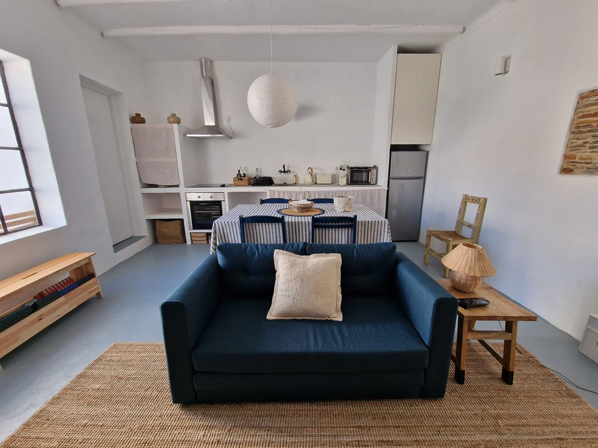 Sala aberta para a cozinha, com sofá azul-escuro, mesa posta com toalha de riscas azuis e brancas, candeeiro de papel e tapete de juta.