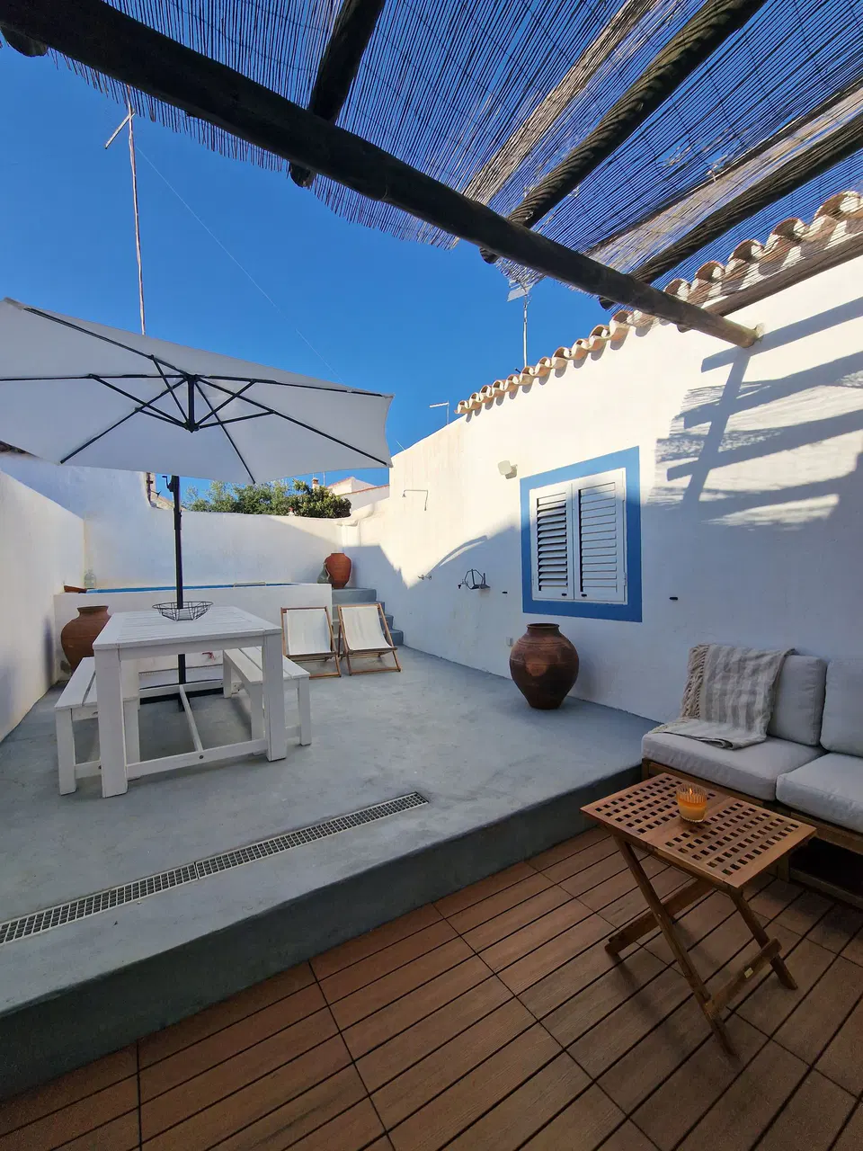 Terraço da casa com mesa branca, guarda-sol e sofá corrido à sombra de uma cobertura de canas, junto a uma portada azul e a talhas de barro.