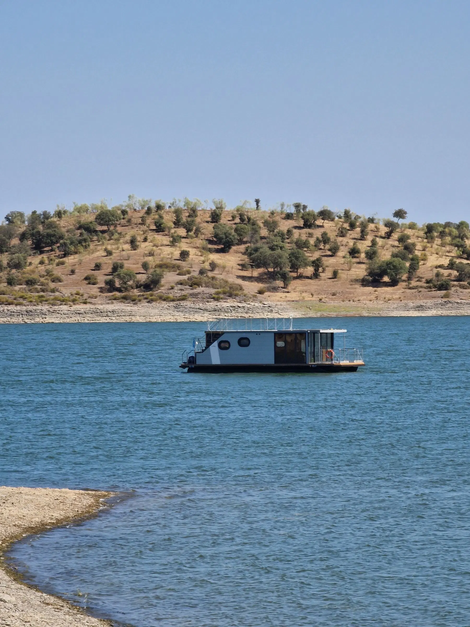 Barco-casa fundeado na água azul do Alqueva, junto a uma encosta seca pontuada de oliveiras.