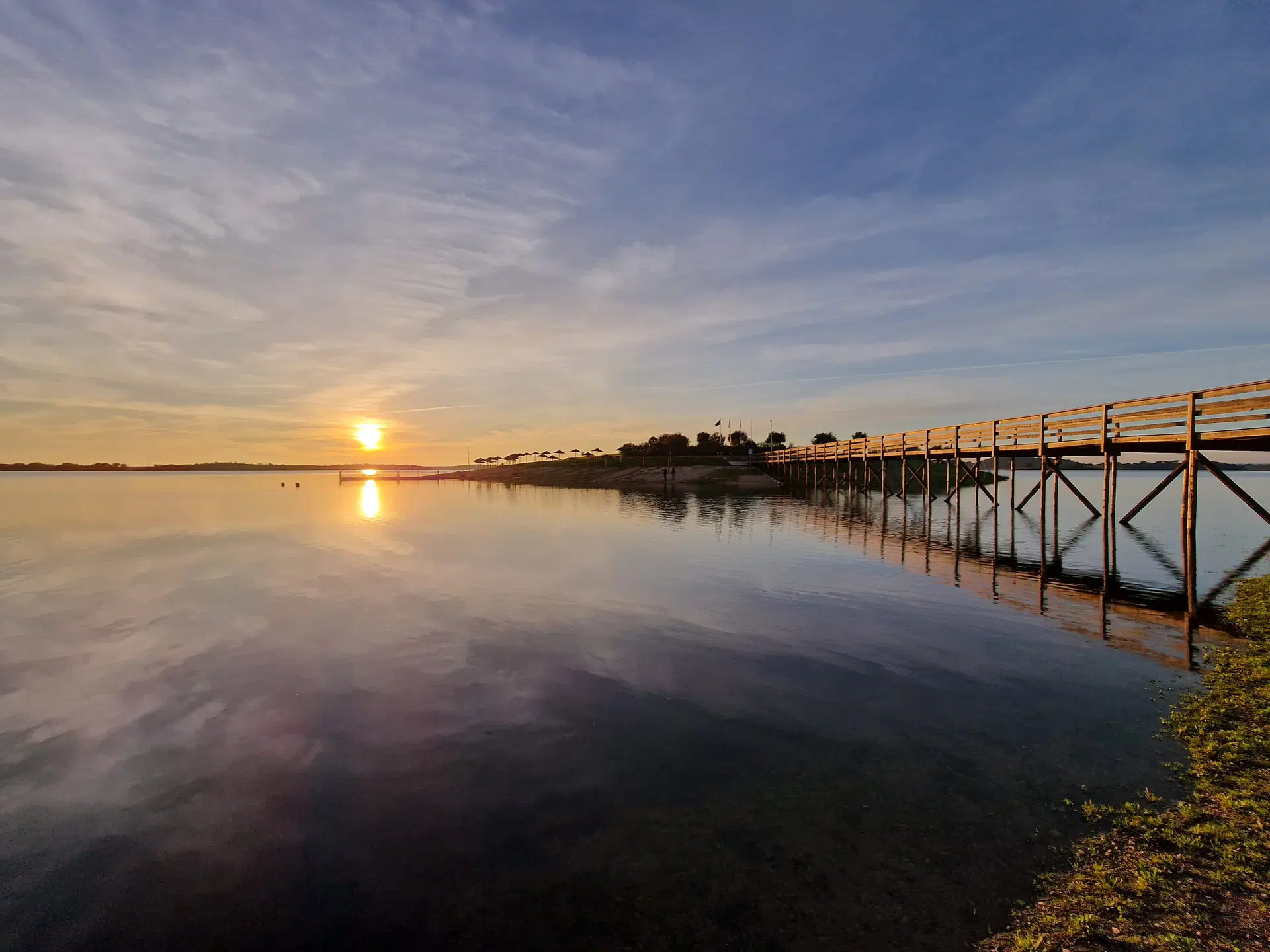 O sol a pôr-se sobre a água na Praia Fluvial de Mourão, com o passadiço iluminado à direita.